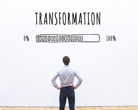 Eine Person steht mit den Händen in den Hüften und blickt auf eine riesige weiße Wand. Darüber prangt „TRANSFORMATION“ neben einem handgezeichneten Fortschrittsbalken, der zu 75 % gefüllt ist und den Fortschritt von 0 % auf 100 % symbolisiert – eine Reise, die an die Weiterentwicklung des Security Service Edge (SSE) erinnert.