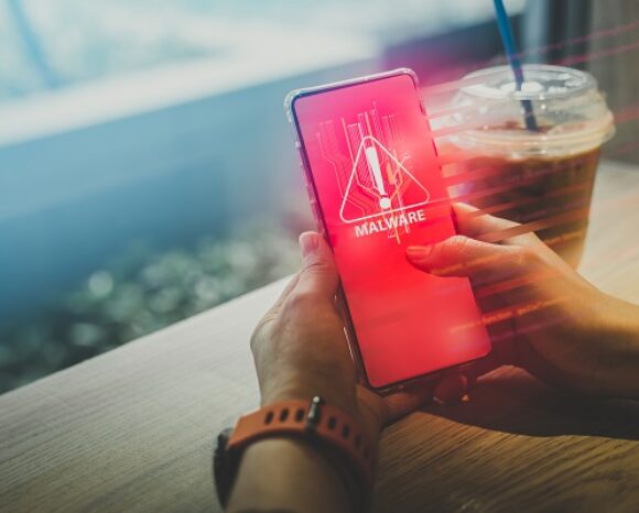 Eine Person hält ein Smartphone in der Hand, auf dem ein roter Bildschirm mit einem Malware-Warnsymbol zu sehen ist, das möglicherweise durch einen E-Mail-Betrug ausgelöst wurde. Ein Getränk mit Strohhalm steht neben ihr auf dem Holztisch. Der Hintergrund ist leicht verschwommen.