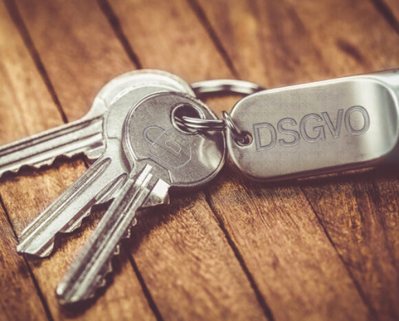Auf einer Holzoberfläche liegt ein Satz silberner Schlüssel, befestigt an einem Schlüsselanhänger mit der Gravur „DS-GVO“.