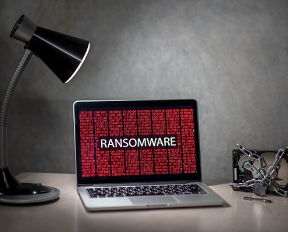 Auf einem Laptop auf dem Schreibtisch steht die bedrohliche weiße Aufschrift „RANSOMWARE“ vor einem roten, verschlüsselten Hintergrund. Daneben, im Schein einer brennenden Schreibtischlampe, liegt eine mit Ketten und einem Vorhängeschloss gesicherte Festplatte – ein deutliches Zeichen für die digitale Verwundbarkeit.
