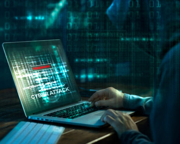 Im schwachen Licht arbeitet eine Person in einem dunklen Kapuzenpullover konzentriert an einem Laptop. Auf dem Bildschirm blinkt „CYBER ATTACK“ vor einem Hintergrund aus Binärcode. Die Szene erinnert an eine düstere Welt der Cyberangriffe und digitalen Intrigen.