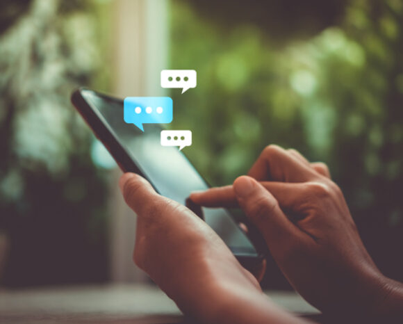 Eine Person hält ein Smartphone in der einen Hand und bedient es mit der anderen. Darüber erscheinen mehrere Chatblasen mit der Aufschrift „Chatkontrolle“ und Auslassungspunkten, die einen laufenden Nachrichtenaustausch signalisieren. Der Hintergrund ist ein sanft verschwommenes Grün.