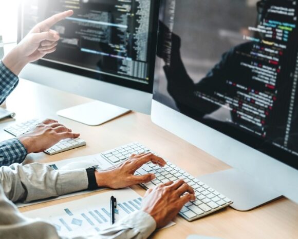 Zwei Personen arbeiten an einem Schreibtisch mit großen Computerbildschirmen zusammen, auf denen Codezeilen angezeigt werden. Einer zeigt auf den Bildschirm, während der andere auf einer Tastatur tippt. Zwischen Papier und Stift verkörpert ihre konzentrierte Teamarbeit den innovativen Ansatz von Schatten-IT in der Softwareentwicklung.