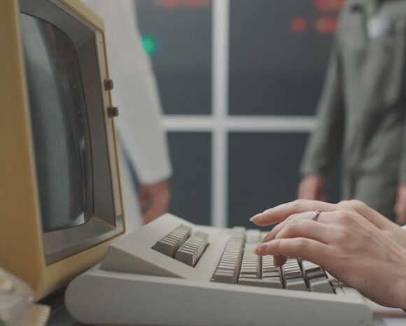 Nahaufnahme von Händen, die auf einer alten Computertastatur tippen. Sie weckt Nostalgie in eine Zeit, bevor Datenschutzverletzungen ein Thema waren. Der klobige Monitor mit seinem beigen Gehäuse summt leise, während im Hintergrund zwei verschwommene Figuren neben einem großen Fenster stehen.