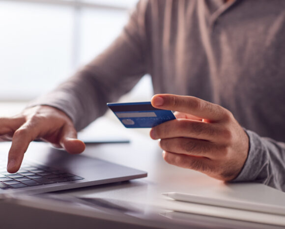 Eine Person in einem grauen Langarmhemd tippt auf einem Laptop und hält dabei eine Kreditkarte in der Hand. Sie ist sich der Betrugsgefahr durch den CEO bewusst. Sie sitzt an einem Schreibtisch, auf dessen Oberfläche zwei Stifte zu sehen sind; der Hintergrund ist leicht verschwommen.