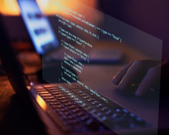 Eine Nahaufnahme eines Laptops, auf dessen Bildschirm Code angezeigt wird, mit einem dezent sichtbaren Microsoft-Logo. Eine Hand ist leicht im Hintergrund zu sehen, was darauf schließen lässt, dass jemand tippt. Reflexionen des Codes schimmern auf der schwach beleuchteten Oberfläche der Laptop-Tastatur.