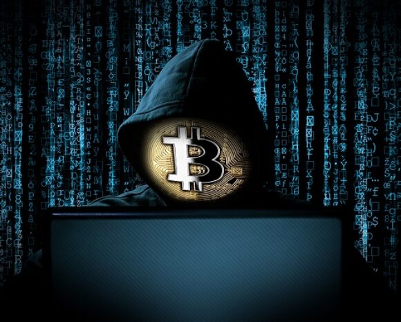 Eine Person in einer Kapuzenjacke sitzt an einem Laptop mit einem großen Bitcoin-Symbol über dem Gesicht, umgeben von digitalem Code, der an Matrix erinnert. Die Szene vermittelt Themen wie Kryptowährung, Hacking und die lauernde Präsenz von Schadsoftware.