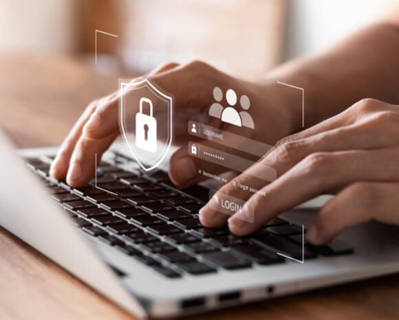 Hände tippen auf einem Laptop, über dem digitale Sicherheitssymbole eingeblendet sind, darunter ein Vorhängeschloss und Benutzeranmeldesymbole, die Konzepte der Cybersicherheit und des Datenschutzes inmitten einer sich entwickelnden Bedrohungslage darstellen.