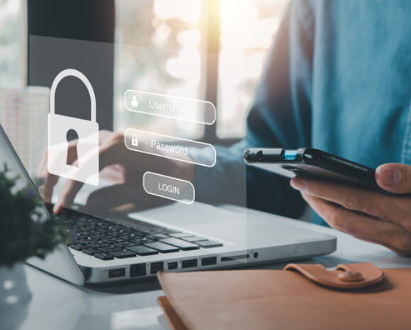Eine Person sitzt mit einem Laptop an einem Schreibtisch und hält ein Smartphone in der Hand. Auf dem Bildschirm erscheint ein schwebender Bildschirm mit Anmeldefeldern für Benutzername und Passwort sowie einem Vorhängeschloss-Symbol – ein Symbol für Online-Sicherheit. Helles Fensterlicht fällt vom Hintergrund herein.