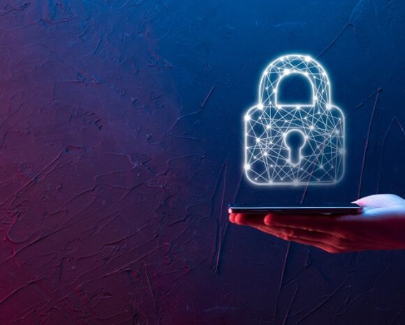 Eine Hand hält ein Smartphone, über dem ein holografisches Vorhängeschloss-Symbol schwebt – ein Symbol für digitale Sicherheit von Kaspersky. Im Hintergrund ist eine strukturierte Wand mit blauen und roten Lichteffekten zu sehen.