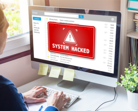 Eine Person sitzt an einem Schreibtisch und starrt mit weit aufgerissenen Augen auf den Computerbildschirm. Auf dem Bildschirm erscheint eine Warnung mit einem rot-weißen Dreiecksymbol und dem Text „SYSTEM GEHACKT“. Trotz ihrer Cybersicherheitskenntnisse überrascht sie dies. Post-its zieren den Monitor; eine Pflanze steht ruhig auf dem Schreibtisch.