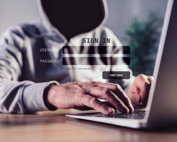 Eine Person im Kapuzenpullover tippt auf einem Laptop. Eine halbtransparente Anmeldeoberfläche zeigt die Felder für Benutzername und Passwort. Ein Link für vergessene Anmeldedaten und eine Schaltfläche „Weiter“ sind ebenfalls sichtbar und runden die Szene der Cyberinteraktion ab.