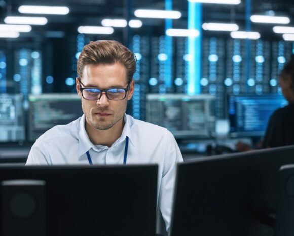Eine Person mit Brille und weißem Hemd konzentriert sich auf Computerbildschirme in einem schwach beleuchteten Büro voller Monitore und Geräte. Eine weitere Figur ist im Hintergrund verschwommen und deutet auf ein Technologiezentrum oder eine Produktionssystemumgebung hin.