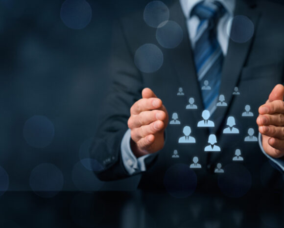 Eine Person im Anzug hält ihre Hände mit holografischen Personensymbolen auseinander, die ein Netzwerk oder Team symbolisieren. Die Szene vermittelt vor dem dunklen Hintergrund ein Gefühl von Identitätsschutz, das durch kreisförmige Lichteffekte verstärkt wird.