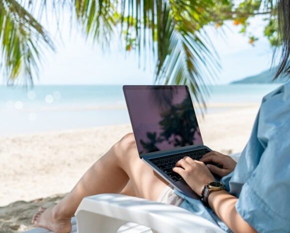 Eine Person sitzt auf einem Stuhl unter Palmenblättern und arbeitet an einem Laptop an einem Sandstrand mit Blick auf das Meer und die fernen Hügel. Die Szene ist sonnig und ruhig und verkörpert die Essenz von „Arbeiten im Urlaub“.