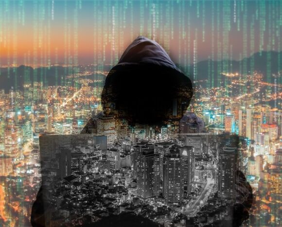 Eine Person in einer Kapuzenjacke sitzt im Schatten verborgen an einem Laptop. Die hellen Lichter der Stadt leuchten, während ein grüner Binärcode, der an REvil erinnert, von oben herabfällt.
