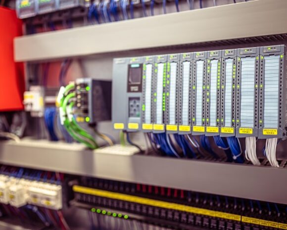 Nahaufnahme eines industriellen Steuerungssystem-Panels mit übersichtlich angeordneter Verkabelung und einer Reihe beschrifteter Module. Links ist eine rote Komponente zu sehen, während mehrere LED-Anzeigen und angeschlossene Kabel den Aufbau vervollständigen.