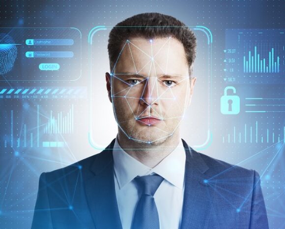 Ein Mann im Anzug steht vor einer digitalen Benutzeroberfläche, die biometrische Daten, Grafiken und Sicherheitssymbole präsentiert. Eine Gesichtserkennungs-Overlay auf seinem Gesicht suggeriert Wachsamkeit gegenüber technologischen Bedrohungen wie Deepfake-Angriffen und unterstreicht die Bedeutung modernster Sicherheitsmaßnahmen.