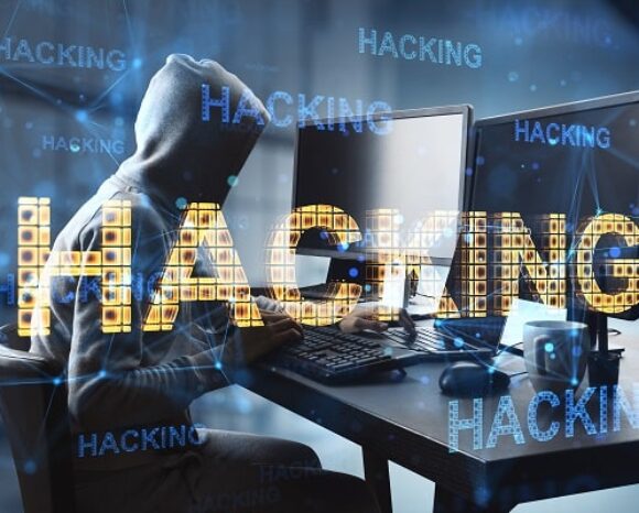 Eine Person in einem Kapuzenpullover sitzt an einem Schreibtisch mit mehreren Computerbildschirmen. Das Wort „HACKING“ ist in fettem, digital anmutendem Text deutlich darübergelegt. Der dunkle Hintergrund zeigt den Begriff „Datenbeute“ und digitale Netzwerkbilder, was das Cyber-Thema verstärkt.