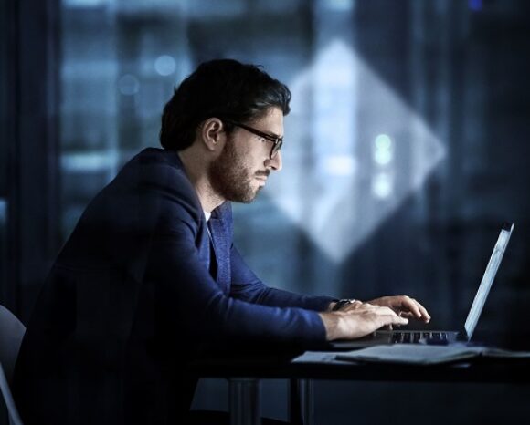 In einem schwach beleuchteten Raum ist ein Mann mit Brille und Anzug tief in seinen Laptop vertieft. Der Schein des Bildschirms hebt sein Gesicht hervor. Vor dem verschwommenen Hintergrund eines eleganten, modernen Interieurs, das durch Glaswände sichtbar ist, stellt er sich der komplexen Herausforderung durch Ransomware-Bedrohungen.