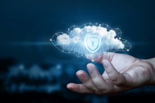 Eine nach oben ausgestreckte Hand hält eine digitale Wolke mit einem Netzwerkmuster. Auf der Wolke ist deutlich ein Schild mit einem Schlüssellochsymbol zu sehen, das für Cloud-Sicherheit steht. Der Hintergrund ist dunkelblau und unscharf.