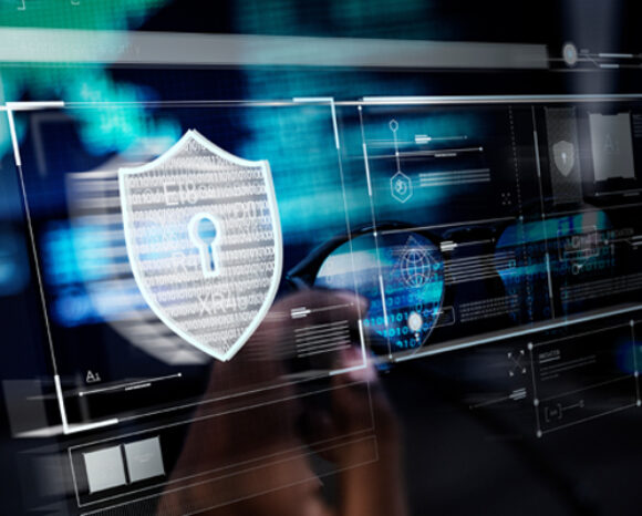 Eine digitale Schnittstelle zeigt ein großes Schildsymbol mit Schlüsselloch und suggeriert damit DS-GVO-konforme Cybersicherheit. Im Hintergrund sind verschiedene Datengraphen und Codezeilen zu sehen, die eine hochtechnologische Sicherheitsumgebung vermitteln.