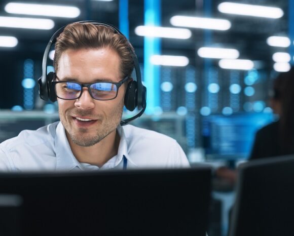 Ein Mann, möglicherweise ein IT-Berater, trägt Brille und Headset und sitzt an einem Computer in einem modernen Büro mit mehreren Bildschirmen und blauem Licht. Er wirkt konzentriert und engagiert. Im Hintergrund arbeitet eine weitere Person am Computer.