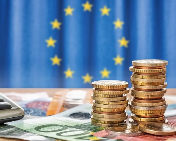 Im Vordergrund liegen Stapel von Euromünzen und verstreuten Banknoten, begleitet von einem Taschenrechner, der präzise Berechnungen symbolisiert. Im unscharfen Hintergrund weht stolz die Flagge der Europäischen Union, die mit ihren gelben Sternen auf blauem Grund ein Bekenntnis zur Digitalisierung verkörpert.