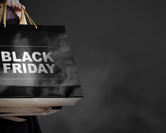 Eine Person hält Einkaufstüten in der Hand. Eine auffällige schwarze Tasche ziert im Schatten des Hintergrunds die Aufschrift „Black Friday“. Drei Seilgriffe stechen hervor, während sie durch die geschäftigen Angebote und Schnäppchen dieses Shopping-Spektakels navigieren.