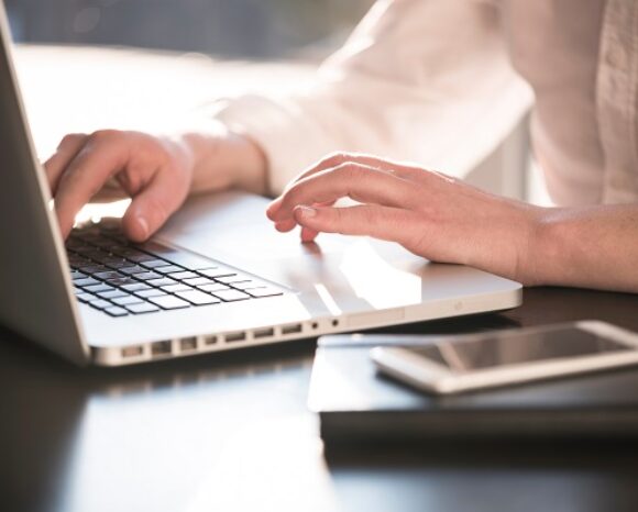 Eine Person im weißen Hemd nutzt einen Laptop auf einem dunklen Tisch. Daneben stehen verschiedene Endgeräte wie ein Smartphone und ein geschlossenes Notebook. Sonnenlicht erhellt die Szene und suggeriert eine Tageslichtumgebung in einem Büro oder einem Heimarbeitsplatz.