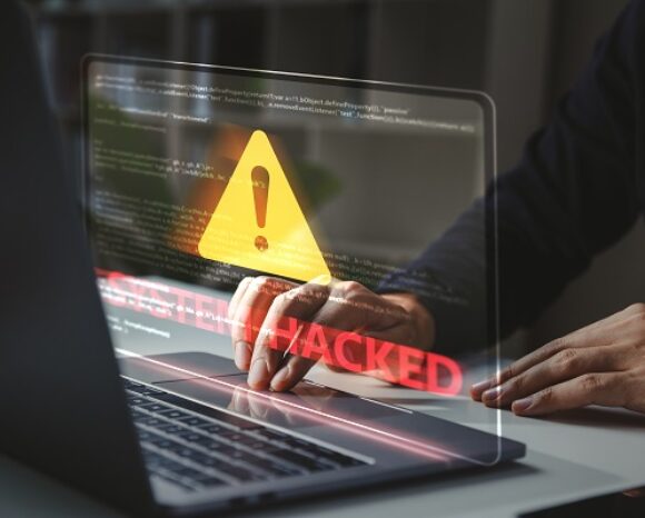 Eine Person tippt auf einem Laptop, auf dem ein Warnschild mit einem Ausrufezeichen angezeigt wird. Auf dem Bildschirm steht in fetten roten Buchstaben „SYSTEM GEHACKT“ – ein Hinweis auf einen Cyberangriff.