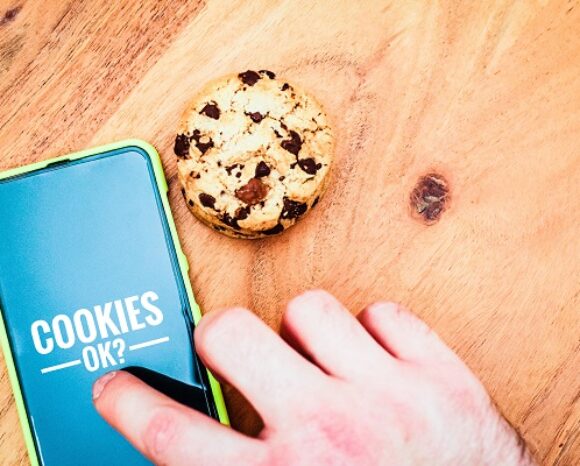Eine Hand schwebt über einem Smartphone-Bildschirm mit der Anzeige „COOKIES OK?“ neben einem Schokoladenkeks, bereit zur Zustimmung zur Nutzung von First-Party-Daten. Das Telefon liegt auf einer Holzoberfläche.