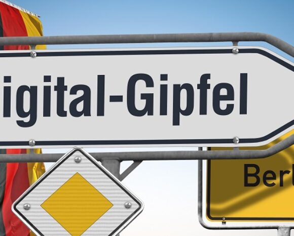 Schild „Digital-Gipfel“ mit Richtungspfeil vor blauem Himmel. Im Hintergrund sind eine Teilansicht der deutschen Flagge und ein Straßenschild mit der Aufschrift „Berlin“ zu sehen.