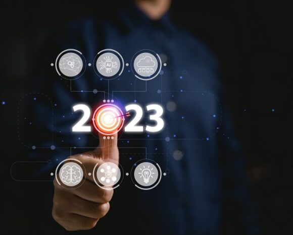 Eine Person drückt einen digitalen „2023“-Knopf, umgeben von futuristischen Symbolen wie einer Wolke, einem Gehirn, einer Glühbirne und Zahnrädern. In dieser düsteren Szene aus leuchtenden Symbolen taucht das Thema der Cybersecurity-Trends 2023 auf: Innovation und digitaler Schutz in einer sicheren Zukunft.