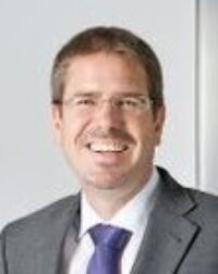 Ein lächelnder Mann in Anzug und Krawatte steht selbstbewusst vor einer metallischen Aufzugstür auf hellem Hintergrund und verkörpert die Widerstandsfähigkeit, die nötig ist, um sich den größten IT-Sicherheitsbedrohungen zu stellen.