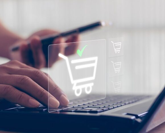 Eine Person bedient einen Laptop, während digitale Einkaufswagen-Symbole auf dem Bildschirm erscheinen. Ein grünes Häkchen signalisiert einen erfolgreichen Kaufabschluss. Gleichzeitig hält sie ein Smartphone in der Hand und liest die Erkenntnisse der BSI-Studie „Onlineshops bergen oft Risiken“.