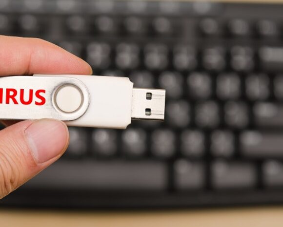 Eine Hand hält einen USB-Stick mit der Aufschrift „VIRUS“ in fetten roten Buchstaben – ein Hinweis auf das Wiederaufleben einer längst vergessenen Bedrohung. Im unscharfen Hintergrund ragt eine Computertastatur hervor, auf der der Satz „Totgeglaubte leben länger: USB-Wurm ist wieder da“ widerhallt.