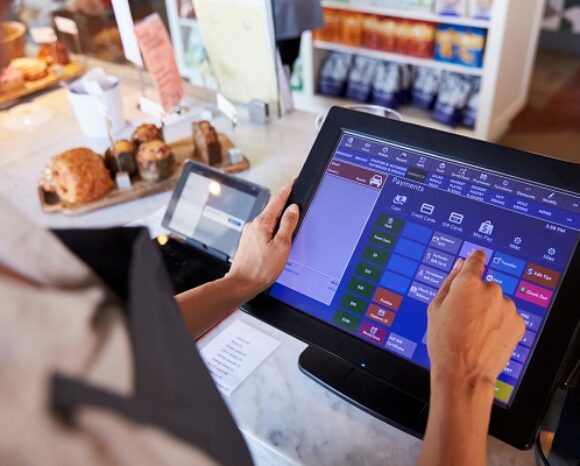 In einem gemütlichen Café bedient eine Person ein elegantes Touchscreen-Kassensystem, während auf dem Bildschirm verschiedene Optionen angezeigt werden. Im Hintergrund locken verführerische Gebäckstücke. Gleichzeitig hat das BSI neue technische Richtlinien zur Sicherung von Public-Key-Infrastrukturen veröffentlicht, um sichere Transaktionen zu gewährleisten.