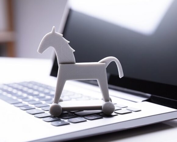 Ein kleines, graues Spielzeugpferd sitzt auf der Tastatur eines aufgeklappten Laptops. Das schlichte Pferd auf Rädern wirkt so, als würde es vor Bedrohungen schützen. Der schwarze Bildschirm spiegelt seine Silhouette wider – ein stiller Wächter der digitalen Welt, der an einen Trojaner aus dem „Monat der Trojaner“ erinnert.