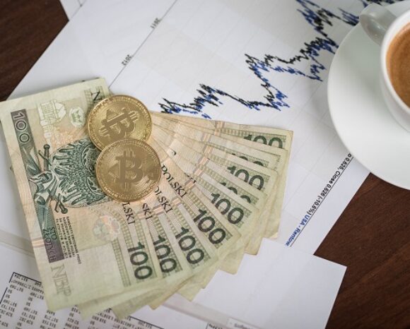 Ein Stapel polnischer Zloty-Scheine und zwei Bitcoin-Token liegen auf einem aufwärts gerichteten Finanzchart und deuten auf einen bullischen Kryptomarkt hin. Daneben steht eine dampfende Tasse Kaffee auf dem Holztisch und rundet das Bild ab.