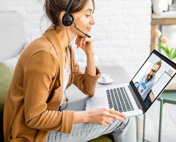 Eine Frau mit Headset sitzt mit einem Laptop auf dem Schoß auf einem grünen Sofa und diskutiert in einem Videoanruf mit einem Mann auf dem Bildschirm die neuesten Sicherheitstrends. Eine Vase mit Tulpen ziert den Tisch neben ihr in dem hellen, modernen Raum.
