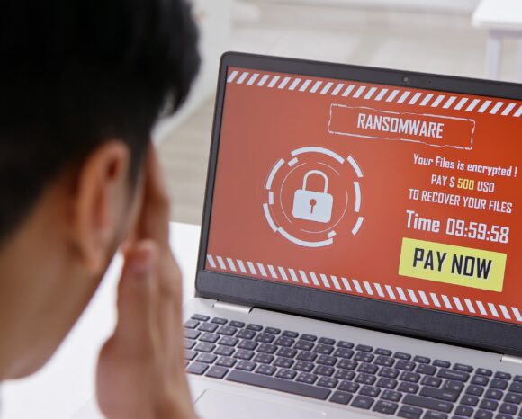 Eine Person starrt auf den Bildschirm eines Laptops, der gerade von einem Ransomware-Boom erfasst wird. Ihre Augen sind auf die bedrohliche Nachricht gerichtet. Die Wiederherstellung der Dateien soll 500 US-Dollar kosten. Ein tickender Timer zeigt 09:59:58 vor rotem Hintergrund, ein Schlosssymbol und einen ominösen „JETZT BEZAHLEN“-Button.