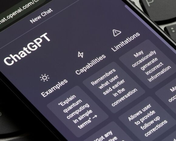 Eine Nahaufnahme eines Smartphone-Bildschirms zeigt die ChatGPT-Webseite mit Abschnitten wie „Beispiele“, „Funktionen“ und „Einschränkungen“, was die Vielseitigkeit unterstreicht. Das Telefon liegt auf einer Laptop-Tastatur. Kommentar: „Sperren von ChatGPT nicht zielführend“ beleuchtet die anhaltenden Diskussionen über Zugriffsbeschränkungen.