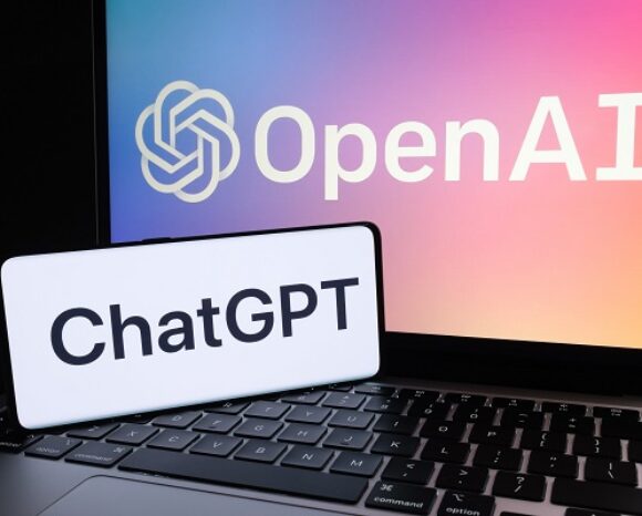 Eine Laptoptastatur mit einem leuchtenden OpenAI-Logo auf dem Bildschirm. Darauf liegt ein Smartphone mit deutlich sichtbarem „ChatGPT“-Schriftzug. Der Hintergrund ist ein bezaubernder Farbverlauf aus Rosa-, Orange- und Blautönen.
