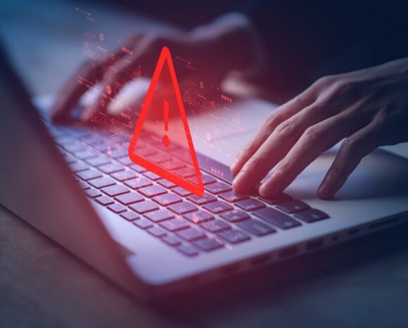 Hände tippen auf einer Laptop-Tastatur. Auf dem Bildschirm erscheint ein rotes Warnsymbol mit Ausrufezeichen, das eine IT-Sicherheitswarnung oder Fehlermeldung signalisiert. Der dunkle Hintergrund lenkt die Aufmerksamkeit auf den ernsten Sicherheitshinweis und die fleißigen Hände.