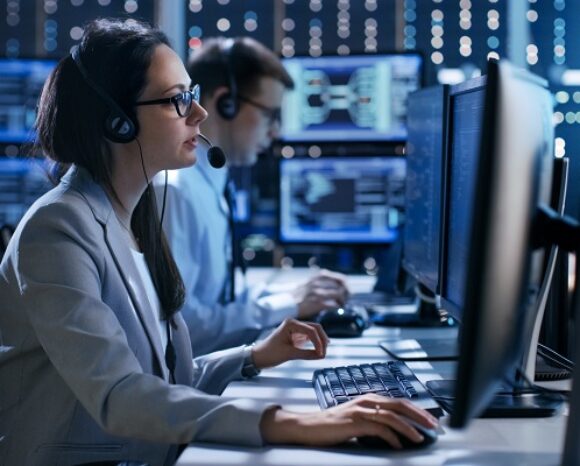 Zwei Personen sitzen an Schreibtischen in einem Büro voller digitaler Datenanzeigen und Schaltkreismuster, die an einen SOC-as-a-Service-Hub erinnern. Sie tragen Headsets, konzentrieren sich auf ihre Aufgaben und sind in dieser modernen Tech-Umgebung von mehreren Computerbildschirmen umgeben.