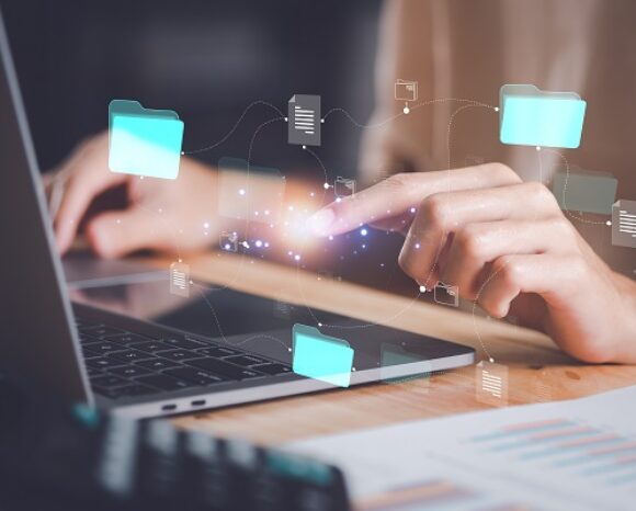 Eine Person interagiert mit einem Laptop und einer digitalen Schnittstelle, auf der leuchtende Symbole von Ordnern und Dokumenten schweben. Ein Diagramm auf Papier deutet auf Datenmanagement oder digitale Dateiorganisation hin, nahtlos unterstützt durch Künstliche Intelligenz.