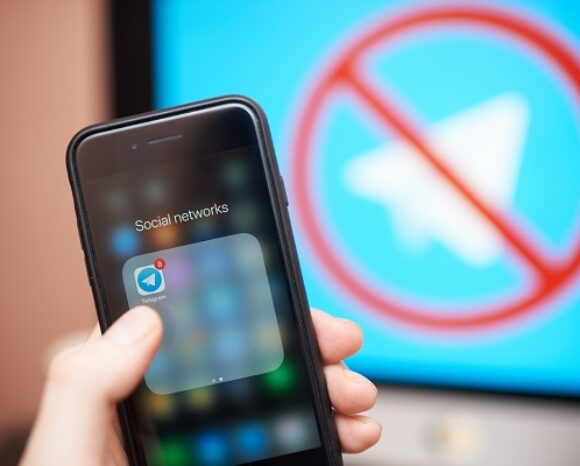 Eine Hand hält ein Smartphone, auf dem ein Ordner „Soziale Netzwerke“ mit dem Telegram-App-Symbol zu sehen ist. Im Hintergrund zeigt ein Bildschirm über dem Telegram-Logo ein rotes Verbotssymbol, das auf einen Telegram: Hinterhältiger Klon hindeutet, der sich direkt vor unseren Augen verbirgt.