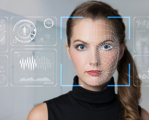 Eine Frau mit ernstem Gesichtsausdruck steht vor einer digitalen Schnittstelle, auf der holografische Grafiken angezeigt werden, darunter biometrische Gesichtserkennungsmuster und Datendiagramme zu Deepfakes. Sie trägt ein schwarzes Oberteil und hat ihr Haar zu einem tiefen Pferdeschwanz gebunden.