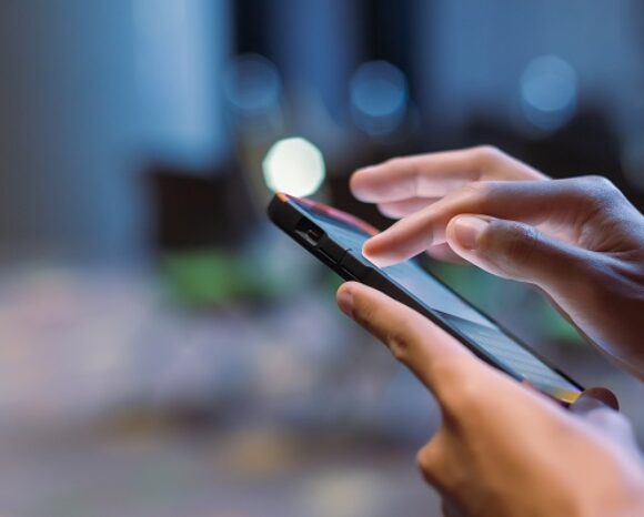 Nahaufnahme von Händen, die ein Smartphone halten, wobei eine Hand auf den Bildschirm tippt. Der Hintergrund ist unscharf und weist sanfte Bokeh-Lichter auf. Dies unterstreicht die Forderung von Digitalcourage nach einem sofortigen Stopp der Chat-Kontrolle nach den jüngsten Erkenntnissen.